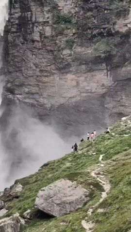 This video shows a waterfall cascading down a rocky cliff. Several people are visible on a grassy slope to the right of the waterfall, walking along a path. The mist from the waterfall creates a cloudy effect around its base.