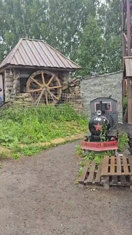 This media file captures an outdoor scene, likely a rural or historical attraction, in Bol'shoi Mashlyak, Russia. The focal point is a rustic wooden structure with a large water wheel, suggesting a historical mill or a replica. Next to it sits a small, decorative model of a steam locomotive, painted black with a red star emblem and red trim, placed on wooden pallets.

The setting is lush and green, with abundant vegetation, including tall trees in the background and overgrown grass and plants around the structures. The path in the foreground is dirt, with some wooden planks forming steps.

In the background, on a grassy slope, a dark-colored horse is visible, along with some farm equipment, including a red tractor and a green vehicle that appears to be a vintage truck or jeep. The sky is overcast, indicating a cloudy or possibly rainy day. The overall atmosphere suggests a quiet, somewhat melancholic day, possibly due to the weather.

A white sign with red Cyrillic text is partially visible on the left side of the frame. The legible text reads "МУЗЕЙ" (Museum) and "1960". This suggests the location is a museum or an exhibition related to the year 1960 or established in that year.

There are no people clearly visible in the frame. The activity appears to be static, showcasing historical artifacts and the natural environment. The presence of the water wheel and the locomotive model hints at an exhibition related to industry, agriculture, or local history.