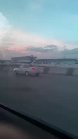 The video, taken from inside a moving vehicle, shows cars driving on a road near a building. The sky is a mix of blue, pink, and white, suggesting dawn or dusk. The building in the background appears to be a terminal. Cars pass by.