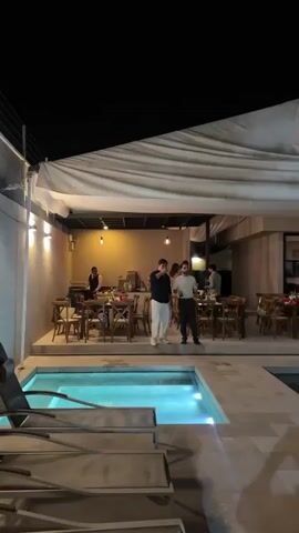 The video shows an evening social gathering by a swimming pool. The pool is rectangular, with blue-lit water. Around the pool area are people conversing and socializing. Some are holding red cups, suggesting they might be drinking. Tables and chairs are visible under a large white awning. The surrounding building appears to be a modern-style house or event venue with large glass doors and a stone accent wall. The atmosphere seems lively and relaxed, consistent with a party or casual get-together.