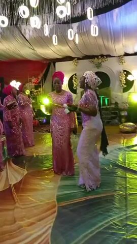 The video shows a group of people, mostly women, dancing at what appears to be a wedding reception or formal event. The women are dressed in matching pink, sequined outfits and headwraps. They dance on a colorfully lit dance floor under a decorated ceiling with hanging lights. There are other guests in the background and photography equipment visible, suggesting the event is being professionally documented. The atmosphere is celebratory and lively.