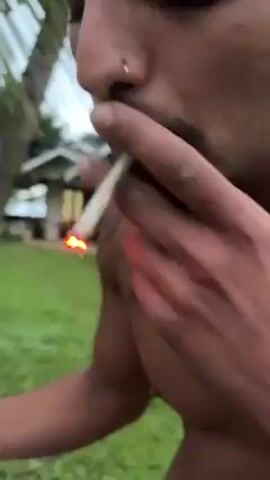 A shirtless man with a nose ring, mustache and tattoos on his chest and arm stands outside in front of a house on a grassy lawn. He looks at the camera and says something in a language other than English. There is a palm tree to his left.
