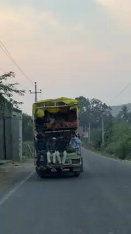 The video shows a small truck driving away on a paved road. The truck has two levels filled with people. The lower level has people sitting on the edge of the truck bed. The upper level has people sitting under a yellow tarp. The road is flanked by trees and power lines. The sky is overcast.