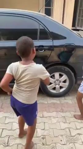 Two young boys are playing in a driveway next to a black car. One boy wears a white t-shirt and blue shorts, while the other wears a blue and green striped shirt and patterned blue and white shorts. They are holding hands and appear to be dancing or spinning around. A girl in a red top and a headscarf is leaning against the car looking bored. The driveway is paved with light-colored bricks. A building with a window is visible in the background.