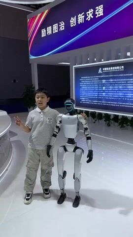 A young boy is standing with his arm around a standing robot. The boy is wearing a striped shirt, khaki pants, and sneakers. He is holding up two fingers in a peace sign. The robot is white and black, with a headpiece that is lit up with a blue light. The robot's arm is around the boy's shoulder. They are in front of a display at a convention. The display features a large screen with Chinese text, and a smaller screen with a black background and white text. There are plants arranged in front of the screens. The floor is white and shiny.