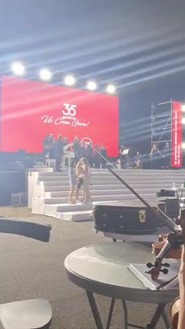 A stage setup for an outdoor event is visible at night. A large red screen dominates the background, displaying "35 Godina Uz Crnu Goru!". On the stage, a group of people, some appearing to be performers or dignitaries, are gathered. A white staircase leads up to the stage. In the foreground, a table holds a violin and its case. The scene is lit by stage lights.