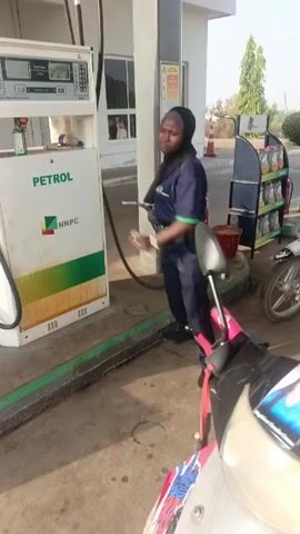A woman wearing a black headscarf and a blue uniform with green trim is filling a white motorcycle with gas at a petrol station. The gas pump is branded "PETROL" and "NNPC." Another man is visible in the background filling another motorcycle. A fire extinguisher is visible near the gas pump. The woman is focused on filling the gas tank. A sticker with a woman's face on it is attached to the motorcycle. The motorcycle has a striped red and black net seat cover.