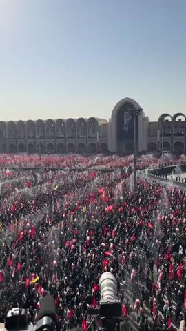 A vast crowd of people, predominantly dressed in black, fills a large outdoor plaza in front of an imposing, arcaded building. Many in the crowd are holding up red and green flags. Water is being sprayed from multiple points into the crowd, creating a misty effect. In the background, a large portrait of a man is visible on the facade of the building. The sky is clear and blue.