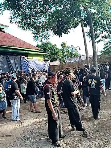 A group of people in black attire are gathered in an open, muddy area, surrounded by trees and buildings. Some individuals are standing, while others appear to be performing some kind of dance or movement in the center. Many of the people are wearing headbands and some of the dancers have decorated clothing. A large tarp is visible in the background.