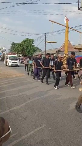 The video shows a procession on a paved road. In the foreground, a person with white and black striped hair is partially visible, with their arm raised. Behind them, a group of people are walking in a line. Several individuals are dressed in military-style camouflage uniforms, while others wear casual clothing. One person is carrying a pole horizontally across their shoulders, supported by two others. There's an ambulance with red and white markings on the left side of the road, and trees and buildings are visible in the background. The sky is clear and appears to be daytime.