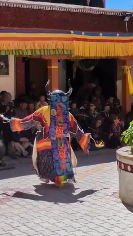A person in a colorful, elaborate costume and mask is dancing in an outdoor courtyard. The costume is multi-layered with a dark blue patterned outer layer, a yellow underlayer, and colorful sashes. The mask has horns and a menacing expression. The dancer is wearing traditional footwear. A crowd of spectators is seated and standing in the background, watching the performance. Behind the dancer, there is a building with a red roof and decorative yellow and red trim. The ground is paved with interlocking stone tiles. The lighting suggests it is daytime.
