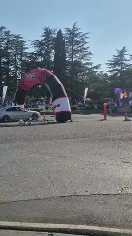 A silver sedan is performing a drift around cones on an asphalt road. The car is positioned in the center of the frame, partially obscured by smoke from its tires. Behind the car, an inflatable archway with a checkered pattern and logos is visible, serving as a start or finish line. Trees line the background, and a building complex is faintly visible in the distance. The sky is clear and blue, indicating a sunny day. Several orange traffic cones are strategically placed on the road to mark the course. The video appears to capture a moment from a motorsports event or demonstration.