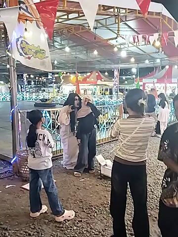 The video depicts an outdoor scene at night in what appears to be a lively amusement park or night market in Sragen, Indonesia. Numerous string lights and illuminated structures create a festive atmosphere. Red and white triangular flags are strung across the area.

In the foreground, several people are gathered. A central figure is dressed as a ghost, wearing a long white gown, long dark hair, and white face paint with exaggerated dark eyes and mouth, reminiscent of a *kuntilanak* or similar Indonesian ghost figure. This ghost character is playfully interacting with another person, a young woman wearing a light-colored hijab and a dark top, who is smiling broadly and being held or embraced by the ghost. Another woman in a striped shirt stands nearby, looking on with a smile, possibly recording the interaction. Further down, another individual is seen lying on the ground, possibly posing or reacting to the ghost. The ground appears to be made of gravel or small stones.

The overall mood is lighthearted and amusing, with laughter and smiles evident among the participants, suggesting a playful scare or photo opportunity rather than genuine fright.