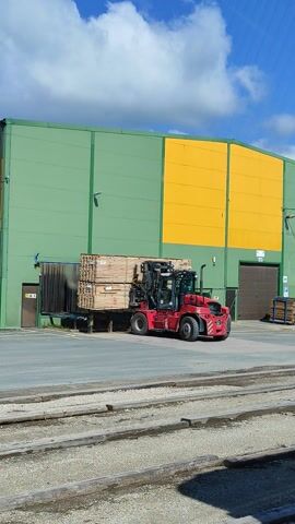 This aerial video, filmed in Verijärve, Estonia, captures a bright, sunny day with scattered clouds. The scene is dominated by a large, industrial building with sections of green and yellow siding. In front of the building, a red forklift is actively engaged in moving materials. The forklift is carrying a large stack of wooden planks, suggesting an operation involving lumber or construction materials. The surrounding area appears to be a logistics or industrial yard, with gravel-covered ground and railway tracks running parallel to the building.

The forklift's movement and the presence of stacked materials indicate an ongoing work operation, likely loading or unloading cargo. The clear skies and abundant sunlight suggest this activity is taking place during daylight hours, possibly mid-morning or afternoon. While no people are clearly visible in the immediate vicinity of the forklift, the operational nature of the scene implies human activity is present. The overall impression is one of industrial productivity in an outdoor setting. No visible text is discernable in the video.