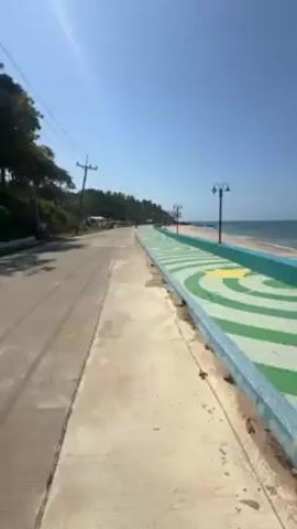 The camera follows a long, paved walkway alongside a road running parallel to a beach. The walkway is decorated with a repeating pattern of concentric green and white curves on a pale blue background, bordered by a light blue wall.  A sandy beach is visible to the right, adjacent to the walkway, with the ocean extending into the distance under a clear blue sky.  Lamp posts are spaced along the edge of the walkway next to the beach. The road to the left has sparse traffic, including a red truck. Trees and vegetation are also present along the left side of the road.  The sunlight is bright.