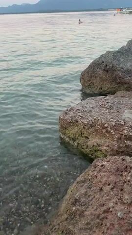 A video displays a serene lakeside scene at dusk. Large, rough rocks line the foreground, with clear water lapping gently against them. The water reveals a gravelly seabed beneath. In the distance, a person is swimming or wading in the lake. Beyond them, mountains rise against a soft, pinkish-purple sky, and a pier with structures and boats extends into the water. The overall atmosphere is peaceful and tranquil.