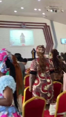 The video shows a gathering of people inside a well-lit building, likely a church or a conference hall. In the foreground, a woman with a vibrant pink and blue headscarf and a matching dress is visible from the side, her hand raised. To her left, a person wearing a teal shirt can be seen. Behind them, other attendees are present, many in traditional African attire. A large projection screen displays an image, possibly of a speaker. A banner reading "FINANCIAL FORTUNE HERITAGE" is prominently displayed on a wall. The seating area is furnished with red chairs. The overall atmosphere appears to be one of a formal or religious event.