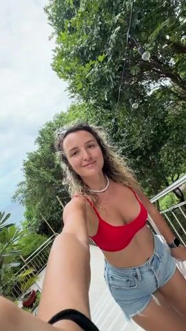 A first-person perspective video shows a woman with curly brown hair and sunglasses pushed up on her head, wearing a red bikini top and denim shorts. She is holding the camera and walking down a white staircase with a metal railing. She smiles at the camera as she descends. The environment appears to be outdoors, with lush green trees and foliage surrounding the staircase. There are also some string lights visible in the background. The railing is made of metal and has some potted plants on the left side. The staircase is wide and seems to lead downwards.