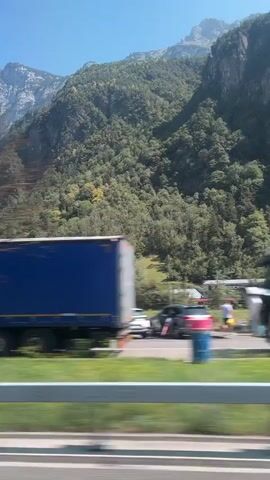 The video shows a view from a vehicle moving along a highway. On the right side of the road, there are large, steep mountains covered in trees. Several vehicles are visible on the highway, including a white truck with a blue trailer, a red truck, cars, and a bright green double-decker bus with a GO FAST sign. A few buildings are scattered along the base of the mountains, and there are guardrails along the side of the highway. The sky is clear and blue.