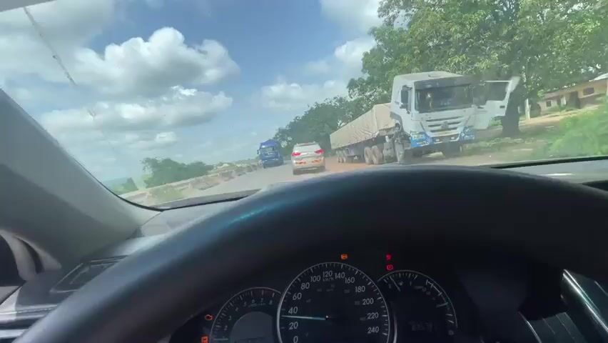 This media file is a first-person perspective video taken from inside a car, looking forward through the windshield. The scene appears to be a highway or road in a rural or semi-rural area, as indicated by the presence of trees and vegetation on the sides. The time of day is daytime, with a bright, partly cloudy sky. The weather seems warm and possibly dusty, given the dirt on the road and the haze in the air.

The primary subjects are vehicles on the road. In the foreground, the steering wheel and dashboard of the car are visible, including the speedometer which shows a speed of approximately 40 km/h. Several large trucks are prominent in the scene, moving in the same direction as the car. One truck, a white cab with a beige tarp-covered trailer, is directly ahead. Further ahead, another truck, a silver DAF tanker truck, is clearly visible. There is also a white SUV driving in front of the tanker truck.

The activity taking place is driving on a road, likely with some traffic. The car is following behind other vehicles, suggesting it might be part of a convoy or encountering a slowdown. The road surface appears to be asphalt but is cracked and uneven in places, with some debris and construction materials visible on the side.

Notable details include the dashboard lights, which appear to be illuminated, suggesting the car is running. There are no people clearly visible inside the car. The emotional tone is neutral, simply documenting the journey.

The location is identified as Aciol, Nigeria. No explicit text is visible in the frame, other than the markings on the vehicle dashboards and road signs that are too indistinct to read.