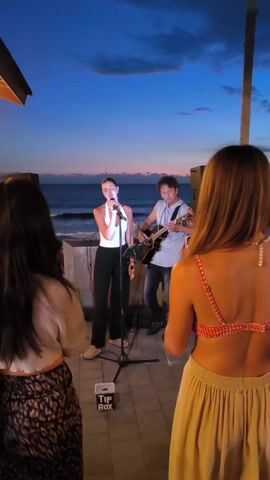 A medium shot shows two female audience members watching a female singer and male guitarist performing at dusk. The singer wears a white top and black pants. The guitarist, slightly behind and to the right of the singer, is wearing a light gray hooded shirt and jeans. They stand on a tiled surface with speakers on either side of them. The singer is using a microphone on a stand, and there's a tip box on the ground in front of her. In the background, the ocean is visible beneath a dusky blue and pink sky. One audience member, to the left of the frame, wears a cropped top and patterned skirt. The other audience member, to the right, wears a red and white bikini top and a tan skirt.