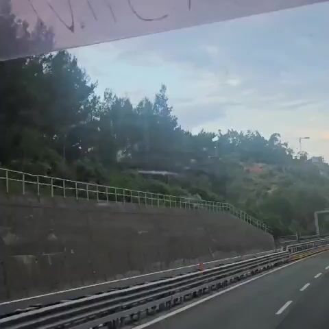 The video presents a view from inside a moving vehicle, looking out through the front or side window. The scene captures a hilly, vegetated landscape under an overcast or partly cloudy sky during daytime.

In the foreground, a multi-lane highway is visible, featuring concrete retaining walls, metal guardrails, and what appear to be railway tracks running parallel to the road. Orange traffic cones are placed alongside the road surface in the initial frames. The hillsides are covered with numerous green trees and dense foliage.

As the vehicle moves forward, a large, historic-looking stone building with a prominent square tower emerges from behind the trees on a distant hilltop. The architecture suggests a church or a castle. The building is light brown or beige, contrasting with the surrounding greenery.

Faint, possibly reversed text is visible on the upper part of the vehicle's window throughout the video. It appears to include the numbers "08" or "80," which could be reflections or markings on the glass. The overall impression is of a journey through a scenic, mountainous region.