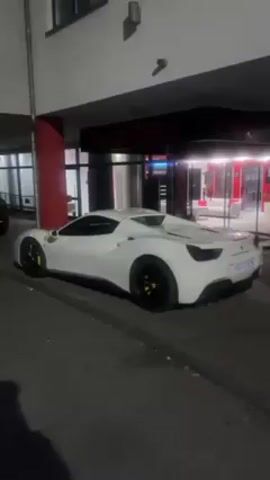 A white Ferrari is parked on the side of a street in front of a building with the word "Donterro" above the entrance. The car is facing left with its back end towards the viewer. It has black wheels and yellow brake calipers. The building is white with a red column to the left of the entrance. The entrance has glass doors and windows that are brightly lit. The street is dark, and the video appears to have been taken at night. There is another car parked further down the street to the left.