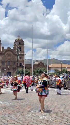 In a vibrant display of culture, a group of traditionally dressed dancers perform on a cobblestone square in front of a large, ornate church with twin towers. The dancers, predominantly young women, wear colorful, layered skirts and elaborate headwear, spinning gracefully to showcase the bright fabric. Several men are also present, wearing ponchos and hats that complement the women's attire. The square is partially covered in confetti, suggesting a festive occasion. A metal barrier separates the performers from a crowd of spectators. The background features additional buildings and a mountainous landscape under a partly cloudy, bright blue sky. Tall metal poles stand in the square, adding vertical lines to the scene.