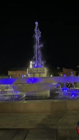 The video shows a fountain at night. The fountain has multiple tiers and is lit with purple lights. Water is spraying from the fountain. In the background, there are buildings and some people. The ground in front of the fountain is paved with bricks.