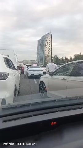 The video shows a traffic jam with several white cars and a large building in the background. There are people standing in the street between the cars, appearing to be interacting with each other or examining the vehicles. The sky is overcast. The timestamp is visible in the bottom left corner of the video.