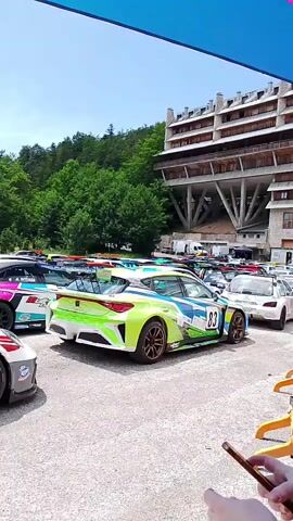 The video shows a parking lot full of race cars on a sunny day. The cars are various colors and designs, with sponsors' logos and racing numbers visible. Some cars are parked close together, while others are spaced out. In the background, there are trees and a building with a distinctive architectural style. A person is partially visible in the foreground, holding a smartphone. The camera pans across the parked cars, providing a view of their exteriors.