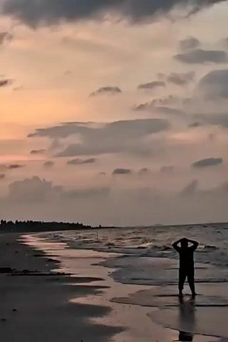 The video shows a silhouetted person standing in the surf on a beach at sunset. The sky is filled with clouds and a gradient of colors from orange to grey. The waves are gently washing onto the shore, and there is a line of trees visible in the background along the coastline. The person has their hands behind their head.