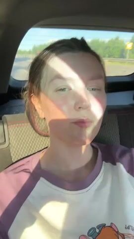 A young woman with light hair is seated in the back of a moving car, looking directly into the camera. She wears a white t-shirt with purple raglan sleeves and a partial graphic design visible on the chest. Strong sunlight streams through the car window, casting a shifting shadow across her face and causing her to squint slightly. She blinks slowly and subtly adjusts her head position while maintaining eye contact with the camera. The car's rear seat and headrest with red stitching are visible, as is a bright outdoor scene with green foliage and a road through the back window.