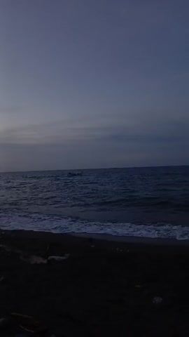 The video shows a dark beach scene. The sky is a muted blue with a layer of gray clouds. The ocean is a deep blue, with small waves coming in towards the shore. The beach itself is dark, appearing to be black sand. There is debris scattered along the shoreline. A faint sound of a vehicle is audible.