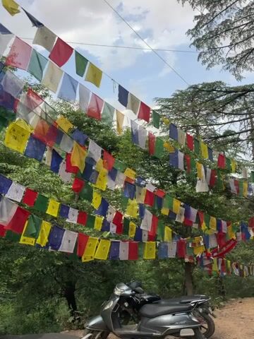 The video shows a road lined with Tibetan prayer flags strung between trees. A gray scooter is parked on the side of the road near a dirt shoulder. The prayer flags are in a variety of colors. A couple of people can be seen walking on the road in the distance. The sky is cloudy.