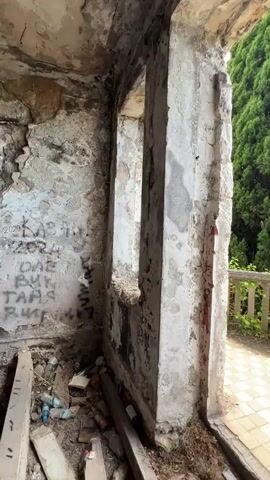 This video shows the interior of a dilapidated building, with a view of a balcony overlooking the sea. The interior is in a state of disrepair, with peeling paint, graffiti on the walls, and debris on the floor, including plastic bottles and wooden planks. The architecture suggests an older building, possibly a historic one, with arched window openings and a stone pillar.

The balcony area is tiled with a checkerboard pattern of beige and reddish-brown tiles. A stone balustrade runs along the edge of the balcony, which is partially overgrown with greenery. Beyond the balustrade, lush green trees, including a very tall, slender evergreen tree, dominate the foreground.

The main focus shifts to the stunning view from the balcony: a vast expanse of turquoise sea under a bright blue sky dotted with white clouds. The horizon line shows the calm sea meeting the sky, with a few distant, indistinct landmasses or structures. The overall impression is one of abandonment and decay within the building, juxtaposed with the timeless beauty of the natural landscape and the sea. The time of day appears to be daytime, likely late morning or early afternoon, given the bright sunlight. The weather seems pleasant, with a clear sky. While no people are visible, the graffiti on the wall suggests human presence at some point. The location cues like the specific architecture and the lush greenery, combined with the coastal view, point towards a Mediterranean or Black Sea region setting, consistent with Abkhazia. The emotion evoked is a mix of melancholy for the decay and awe at the natural beauty. There is visible graffiti on the left wall of the interior, but the text is largely illegible.