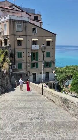 A couple stands on stone steps, facing away from the camera, with the woman in a red dress and the man in a white shirt and shorts. They are holding hands and appear to be interacting. Other people are visible further up the steps and to the left. Behind them is a multi-story building with green shutters and balconies, overlooking the sea. The sky is clear and blue, suggesting a sunny day. The scene appears to be a scenic viewpoint in a coastal town.