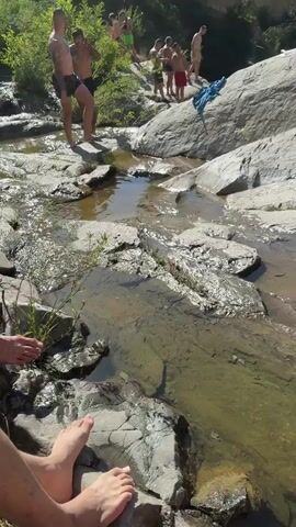 The video shows a group of people at a rocky riverbank on a sunny day. In the foreground, a person's feet are visible, resting on rocks. The middle ground features a shallow stream flowing over rocks, with several individuals gathered on the bank. One man in black shorts is looking down, possibly at a bag. A woman in a blue bikini is adjusting her clothing. Another man in red shorts is holding a phone. Other men are standing around, some in swimwear. The background consists of a steep, vegetated hillside with some red structures visible at the top. A blue towel or piece of clothing is draped over a large rock. The overall scene depicts a casual outdoor gathering near water.