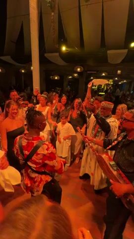 The video captures a lively celebration indoors. A diverse group of people are dancing and enjoying themselves. Some individuals are holding up their phones, likely recording the event. A group of musicians play various instruments, including a small drum, a three-stringed instrument, and percussion. One musician is wearing a traditional white garment with embroidery, while another is wearing a shirt decorated with sequins. The room is decorated with woven lampshades, white draping, and foliage. The lighting is warm and inviting, and there's a partial view of a bar area in the background. The overall atmosphere is festive and energetic.