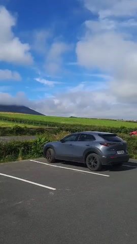 This media file captures a scenic view from a parking lot in Pier Road, Ireland. The foreground is dominated by the asphalt of the parking lot, marked with white lines for parking spaces. A dark gray car is parked in one of the spaces. Lush green hedges border the parking lot, separating it from a vast, rolling green field.

In the middle ground, the field is neatly tilled, with visible rows of crops, possibly grass or hay, stretching towards a distant hill. The hill is partially shrouded in soft, white clouds, adding a dramatic and ethereal quality to the landscape. The sky above is a vibrant blue, dotted with fluffy white clouds that catch the sunlight. The overall impression is one of a beautiful, natural setting bathed in good weather, likely midday or early afternoon given the bright sunlight. There are no people visible in the media. The scene evokes a sense of tranquility and the beauty of the Irish countryside. No visible text is present.