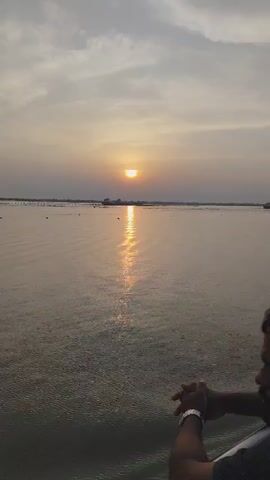 A man stands by a railing watching the sun setting over a large body of water. The sky is filled with clouds, and the water reflects the sun's light. A ship is visible on the horizon. The man is wearing a watch.