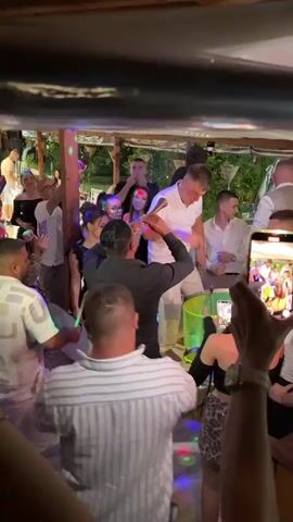 A group of people are gathered in an outdoor space, likely a venue with a covered roof, illuminated by colorful disco lights. In the center of the frame, a man in a black shirt is playing a wind instrument, possibly a clarinet or a zurna, while another man in a white t-shirt and pants dances energetically. Several other individuals are visible, some dancing, some watching, and a few are holding up smartphones to record the scene. One person in the foreground is holding a phone up, capturing a video of the event. The atmosphere appears festive and lively, suggesting a party or celebration. The environment includes wooden structures and foliage, indicating an outdoor setting.