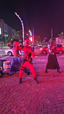 Here's a description of the video:

Three individuals dressed as superheroes are performing a dance in an urban environment at night. The person in the middle is dressed as Deadpool, while the figures to the left and right resemble Spider-Man variations. The Spider-Man on the left sports a cowboy hat and blue pants, whereas the one on the right is dressed in black. They all move in sync. The background includes city lights, vehicles, and modern architecture. The area is illuminated with red light. A drum sits next to the figure on the left. The scene is set on a brick-paved area near what appears to be a street.