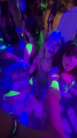 The video begins with a close-up of two young women with lights flashing around them, they are smiling and posing for a selfie. The camera then pans out to show they are in a club or party with many other people dancing and enjoying themselves. There are bright blue and green lights flashing in the background, creating a dynamic and energetic atmosphere. The video continues with shots of the dancing crowd, the flashing lights, and the two women continuing to enjoy the party. Some of the shots are blurry due to the fast movement and low light conditions. The overall impression is a fun and lively party scene.