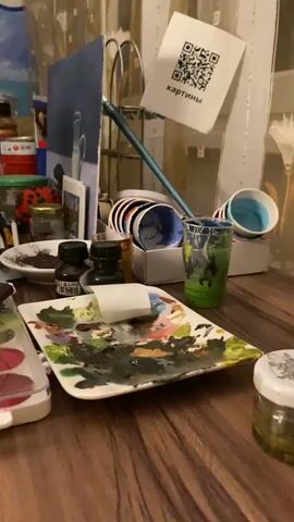 The video shows a close-up, static shot of an artist's cluttered desk. The focal point is a messy palette with dried paint in various colors, including green, brown, pink, and white. Next to the palette are two small, dark bottles, possibly ink or paint. A roll of white paper or cloth is placed on the palette. In the background, there are stacks of small cups or containers with colorful rims, and a larger container holding brushes and other art supplies. A painting or canvas is propped up in the background. A QR code printed on a white piece of paper is attached to a stand, with the Russian word "картины" (paintings) written below it. Other items visible include jars, tubes of paint, and a cigarette pack. The lighting is dim, creating a somewhat moody atmosphere.