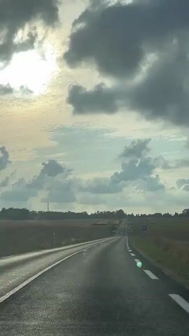 This is a first-person perspective video of a car driving on a paved road. The road is empty except for a few white dashed lines indicating lanes. The sky is cloudy with patches of sunlight breaking through. The landscape on either side of the road appears to be agricultural fields, with some trees and power lines visible in the distance. There is a blue sign with a white "P" on it on the right side of the road.