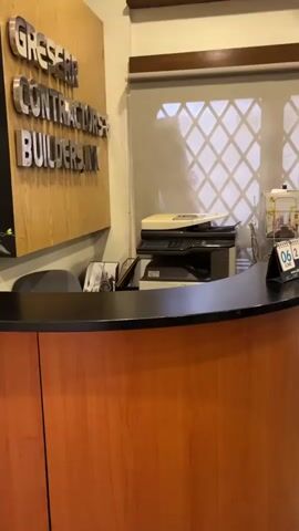 The video opens with a close-up of a reception desk with a wooden front and a black countertop. A sign on the wall behind the desk reads "GRESSER CONTRACTORS BUILDERS INC". To the right of the sign is a copier and a window with blinds. The camera then pans to the right, revealing a light green couch covered with a quilted blanket, a wooden floor, a rocking chair, and a dark wood coffee table with a small figurine on it. The room appears to be an office or waiting area.