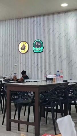 The video shows the interior of a restaurant with tables and chairs. Two neon signs are visible on the wall. One is circular with a yellow background and an image of what appears to be a cup. The other is rectangular with a green background and shows an image of two people. There are several tables with chairs. On one table, there are bottles of water. A young person wearing a black shirt and head covering is sitting at a table, looking down at something in their hands. Another person, partially visible, is sitting at a table to the left. The walls are decorated with a striped pattern.