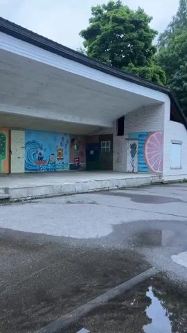 The video pans across an outdoor stage area with amphitheater-style seating. The stage is covered and has a mural on the back wall. The seating consists of rows of wooden benches on metal legs. The ground is paved and wet, with puddles visible. Trees and a grassy field are in the background. The sky is overcast.