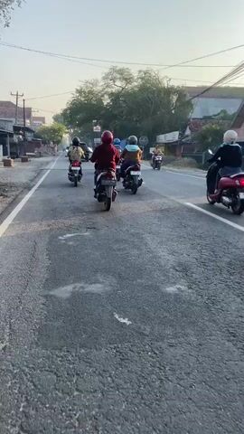 The video shows a group of motorcyclists riding down a paved road. The camera is positioned behind the riders, capturing their backs as they move away. The road appears to be in a semi-urban or rural setting, with trees and buildings visible on either side. Sunlight suggests it is daytime, likely morning or late afternoon, due to the low angle of the light and the warm tones in the sky. Most of the riders are wearing helmets, and some are carrying backpacks. The motorcyclists are riding in a staggered formation, spread across the width of the road. There are at least seven visible motorcycles. One motorcycle in the foreground has a license plate that reads "K 3179 BAX". Further down the road, a white van is visible. The road surface is worn and cracked in places.